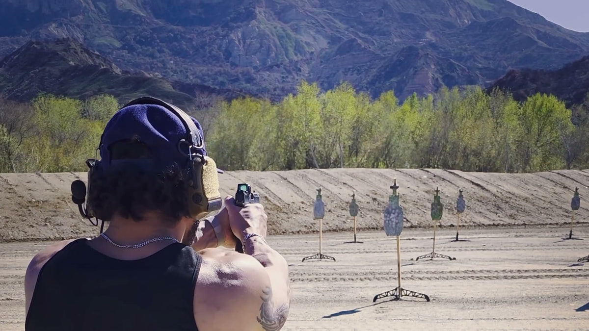 A 2ATG shooter seen from behind, firing a pistol downrange at steel targets with mountains beyond