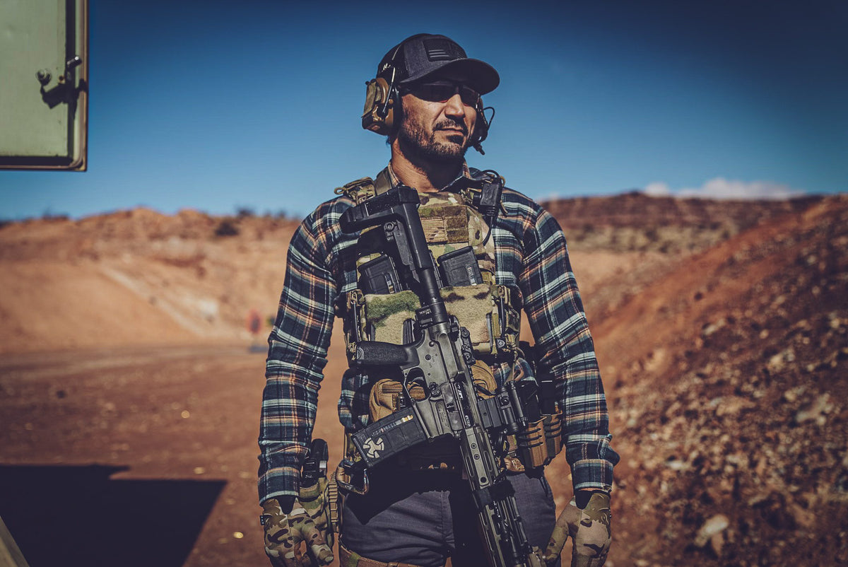A shooter in a chest rig and ear protection carrying a rifle across red desert ground.